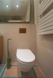 Detalle de inodoro suspendido con pulsador en el mismo acabado que la grifería, sobre pared de cerámica beige, con revestimiento verde y suelo en varios tonos al fondo en el Proyecto Mallorca.